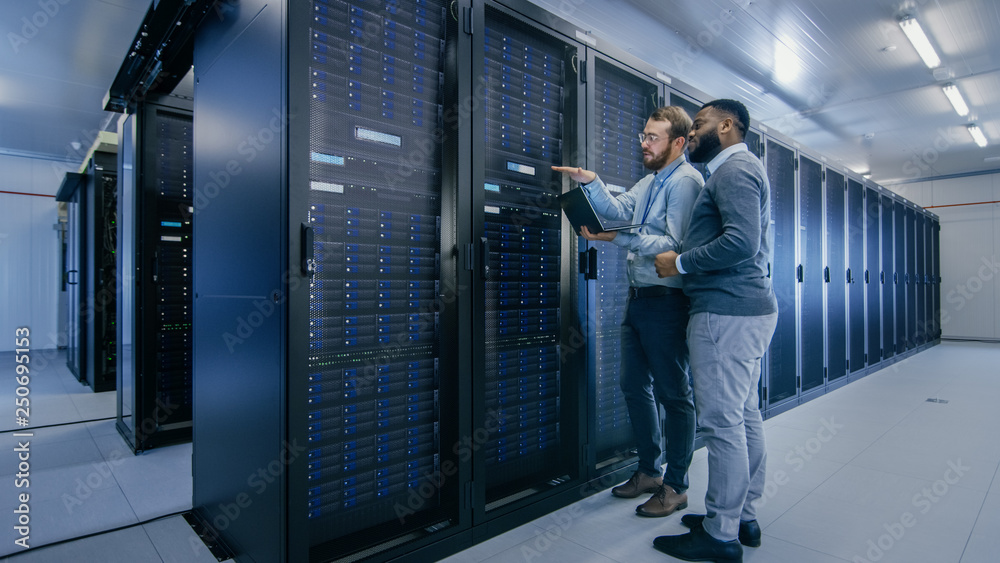 Poster Bearded IT Technician in Glasses with a Laptop Computer and Black Male Engineer Colleague are Talking in Data Center while Working Next to Server Racks. Running Diagnostics or Doing Maintenance Work. 