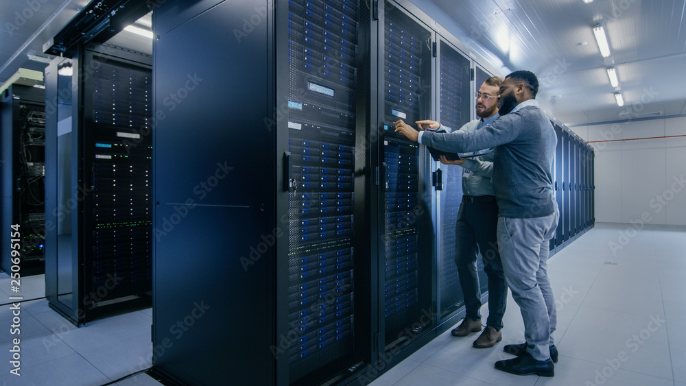 Aufkleber Bearded IT Technician in Glasses with a Laptop Computer and Black Male Engineer Colleague are Talking in Data Center while Working Next to Server Racks. Running Diagnostics or Doing Maintenance Work. 