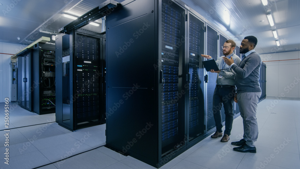 Aufkleber Bearded IT Technician in Glasses with a Laptop Computer and Black Male Engineer Colleague are Talking in Data Center while Working Next to Server Racks. Running Diagnostics or Doing Maintenance Work. 