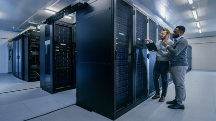 Bearded IT Technician in Glasses with a Laptop Computer and Black Male Engineer Colleague are Talking in Data Center while Working Next to Server Racks. Running Diagnostics or Doing Maintenance Work. 