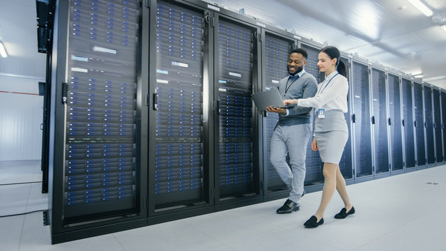 Black IT Technician with a Laptop Computer Gives a Tour to a Young Intern. They Talk in Data Center while Walking Next to Server Racks. Running Diagnostics or Doing Maintenance Work. 