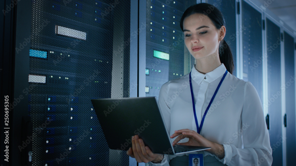 Poster Confident and Beautiful Female IT Engineer is Working on Laptop in Data Center while Standing Before Server Rack. Running Diagnostics or Doing Maintenance Work.