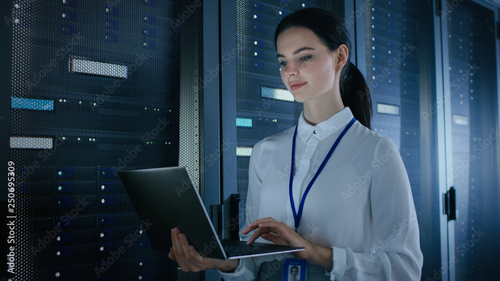 Poster confident and beautiful female it engineer is working on laptop in data center while standing before