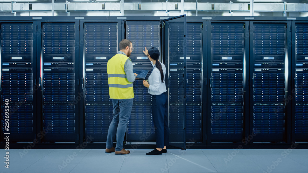 Poster Bearded IT Admin in Glasses and High Visibility Vest with a Laptop Computer and Young Technician Colleague Talking in Data Center while Standing Next to Server Racks Running Diagnostics or Maintenance