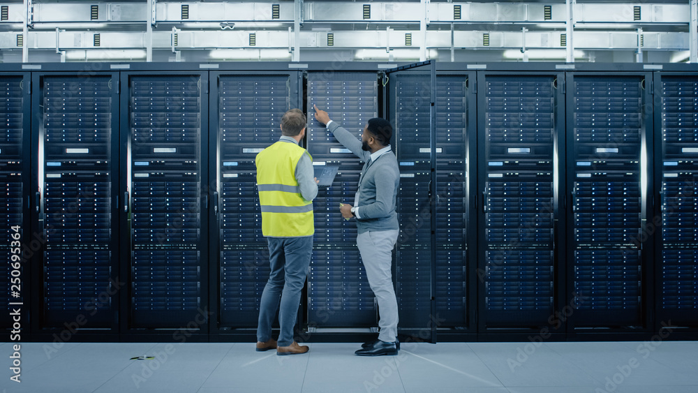 Poster Bearded IT Engineer in Glasses and High Visibility Vest with a Laptop Computer and Black Technician Colleague Talking in Data Center while Standing Next to Server Racks. Running Maintenance. 