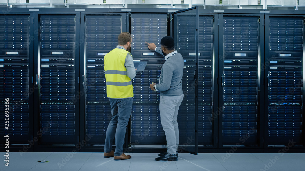 Poster Bearded IT Engineer in Glasses and High Visibility Vest with a Laptop Computer and Black Technician Colleague Talking in Data Center while Standing Next to Server Racks. Running Diagnostics