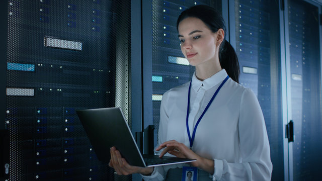 Confident and Beautiful Female IT Engineer is Working on Laptop in Data Center while Standing Before Server Rack. Running Diagnostics or Doing Maintenance Work.