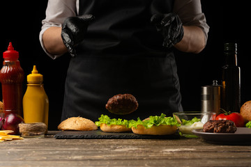 Chef puts beef patty on a burger loaf, against the background of the ingredients. Delicious and harmful food, fast food, homemade recipes, restaurant, catering, recipe book