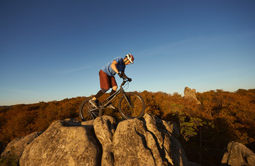 Young male cyclist balancing on trial bicycle on top of big boulder, rider making acrobatic trick on summer evening, blue sky and forest on background. Concept of extreme sport active lifestyle