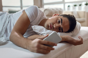 Young sad man lying on bed and reading text message on mobile phone.