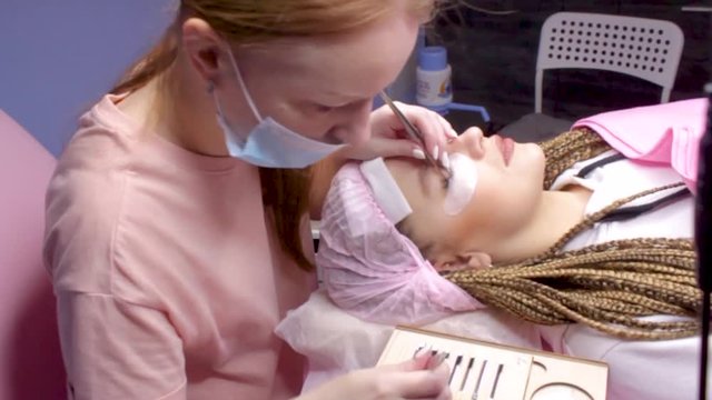 Eyelash Extension Procedure. Woman Eye with Long false Eyelashes. Girl cient lies in beauty salon. Eyelash wizard beautician with tweezers.