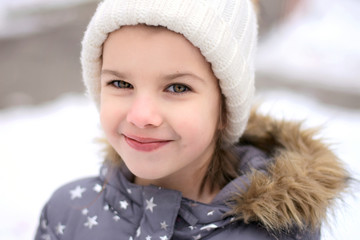 Portrait of smiling beautiful caucasian girl in beige knitted hat and gray coat on blurred winter background. Green eyed girl with a sly happy look looks straight in snow winter park