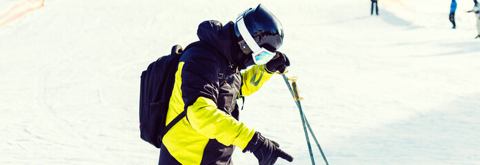 Skier holding ski and looking at beautiful snow covered mountains. Young man thinking and looking snowy mountain with copy space. Side view of winter skier guy with mask holding ski equipment outdoor.