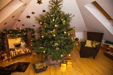 Christmas decorations in the studio, big natural fir tree with golden balls, bows and snowflakes, wooden fireplace and candles on it and brown cozy armchair