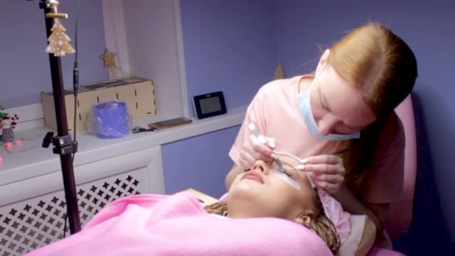Eyelash Extension Procedure. Woman Eye with Long false Eyelashes. Girl cient lies in beauty salon. Eyelash wizard beautician with tweezers.