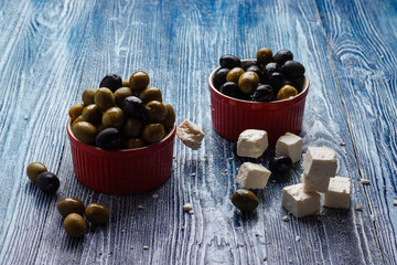 Green and black olives in a red cup, with diced feta (white cheese), on a blue wooden table
