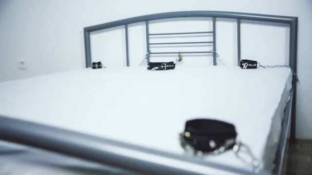 BDSM fetish bed with cuffs and chains. Jib shot from behind the bed frame to uncover cuffs and collar on a white bed.