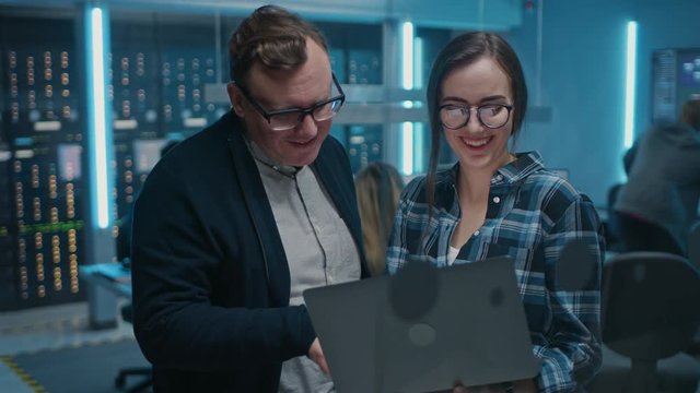 Male IT Engineer and Female IT Programmer Talking, Holding Laptop. In the Background Technical Department with Server Racks and People Working. Concept: Artificial Intelligence, Machine Learning