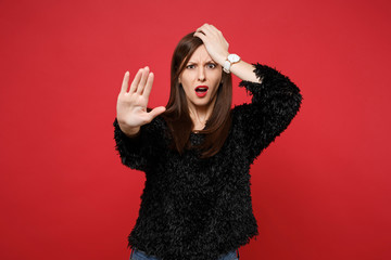 Perplexed young woman in black fur sweater putting hand on forehead, showing stop gesture with palm isolated on bright red background. People sincere emotions, lifestyle concept. Mock up copy space.