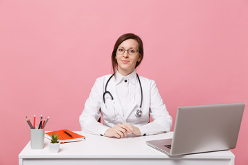 Beautiful female doctor sits at desk works on computer with medical document in hospital isolated on pastel pink wall background. Woman in medical gown glasses stethoscope. Healthcare medicine concept