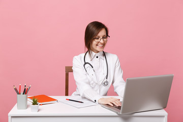 Beautiful female doctor sits at desk works on computer with medical document in hospital isolated on pastel pink wall background. Woman in medical gown glasses stethoscope. Healthcare medicine concept