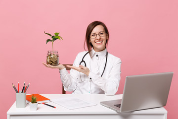 Female doctor sit at desk work on computer with medical document hold coins in hospital isolated on pastel pink wall background. Woman in medical gown glasses stethoscope. Healthcare medicine concept.