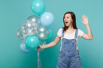 Fototapeta na wymiar Surprised girl looking aside waving greeting with hand as notices someone celebrating hold colorful air balloons isolated on blue turquoise background. Birthday holiday party, people emotions concept.