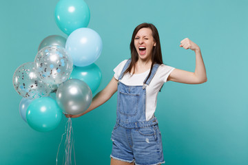 Crazy funny screaming young woman blinking, showing biceps, muscles celebrating, holding colorful air balloons isolated on blue turquoise background. Birthday holiday party, people emotions concept.