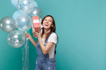 Laughing young girl hold red striped present box with gift ribbon celebrating with colorful air balloons isolated on blue turquoise background. Valentine's Day, Women's Day, birthday, holiday concept.