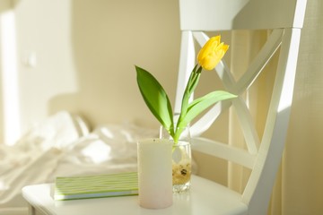 Yellow tulip flower in glass with water on white chair in bedroom interior, sunny spring day