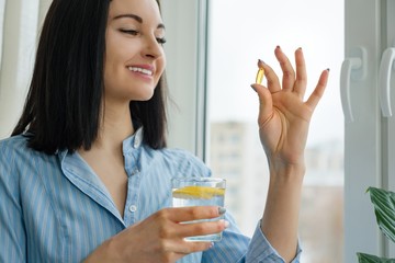 Woman takes pill with omega-3 and holding glass of fresh water with lemon. Morning picture of house, near the window. Vitamin D, E, fish oil capsules. Nutrition, healthy eating, lifestyle