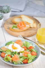 Green salad with lightly salted salmon. On plate with salad toast with poached egg. In the frame of a glass with juice and a plate of bruscheta. Light background. Close-up. Vertical frame orientation.
