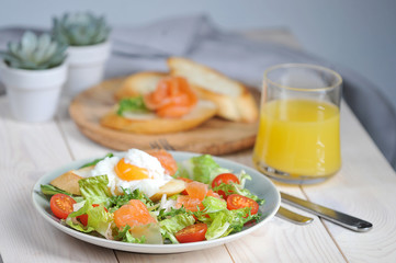 Green salad with lightly salted salmon. On a plate with salad toast with poached egg. In the frame of a glass with orange juice and a plate with bruscheta. Light background. Close-up. 