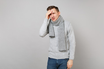 Tired upset young man in gray sweater, scarf having headache, put hand on head isolated on grey wall background. Healthy lifestyle, ill sick disease treatment cold season concept. Mock up copy space.