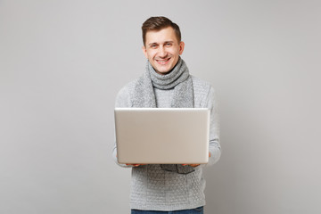 Smiling young man in gray sweater, scarf working on laptop pc computer isolated on grey background in studio. Healthy lifestyle, online treatment consulting, cold season concept. Mock up copy space.