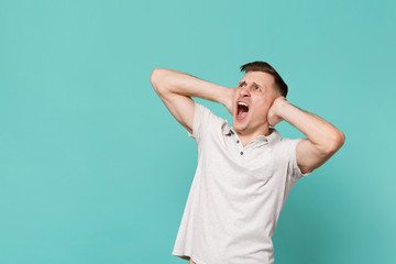 Shocked screaming young man in casual clothes looking up aside covering ears with hands isolated on blue turquoise background in studio. People sincere emotions, lifestyle concept. Mock up copy space.