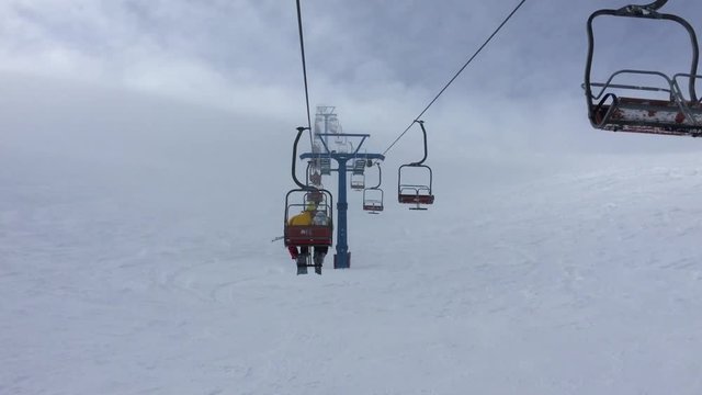 Ski chairlift at a ski resort filmed during a strong wind surrounded by a large number of skiers and snowboarders