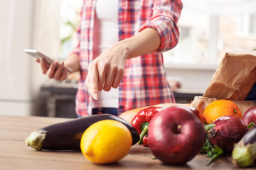 Young girl at kitchen healthy lifestyle standing holding smartphone pointing at bag with products close-up on table