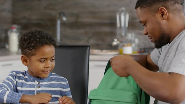Close up of little African boy finishing drawing, giving pencil and notebook to his father packing school bag, and then helping boy putting it on. Little boy going away and his father waving to him