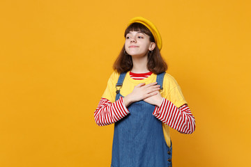 Portrait of pleasant girl teenager in french beret, denim sundress keeping hands on chest, heart isolated on yellow background in studio. People sincere emotions lifestyle concept. Mock up copy space.