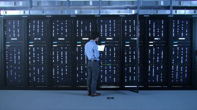 IT Specialist Standing In front of Server Racks with Laptop, He Activates Data Center with a Touch Gesture. Turning On New Data Center.