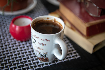 Cup of coffee on table. Old books, glasses, small candle holder and cactuses in the background.