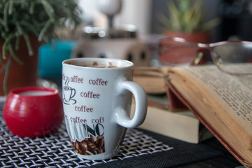 Cup of coffee on table. Old books, glasses, small candle holder and cactuses in the background.
