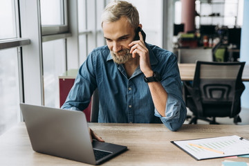 The working process. Stylish guy working in the laptop and talking on the smartphone during the work process in his office