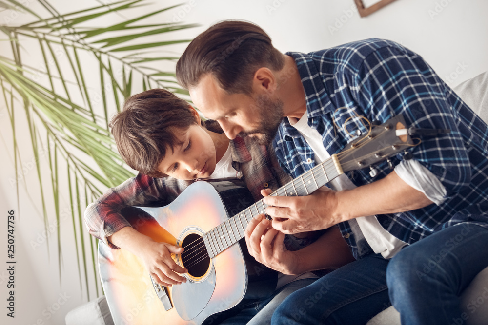 Wall mural father and little son at home sitting on sofa boy with guitar dad showing chords thoughtful