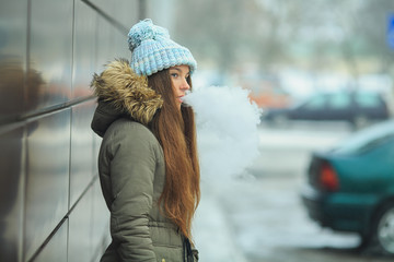 Vape teenager. Young pretty white girl in blue cap is smoking an electronic cigarette on the street in the winter. Bad habit.