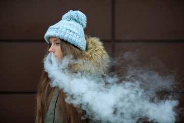 Vape teenager. Young pretty white girl in blue cap is smoking an electronic cigarette opposite modern brown background on the street in the winter. Bad habit. Close up.