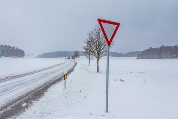 Leere glatte Winter Straße mit Schnee und Vorfahrt achten Schild. Die Fahrbahn ist weiß glatt verschneit. Vorsicht vor Unfall durch Schneesturm und schlechte Sicht. Wetter zur Weihnachten Jahreszeit