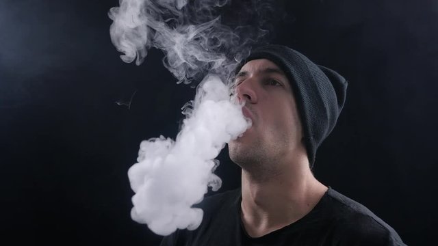young man vaper exhaling big clouds of smoke with e-cigarette vape on black background in slow motion