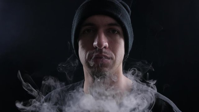 young man vaper exhaling big clouds of smoke with e-cigarette vape on black background in slow motion